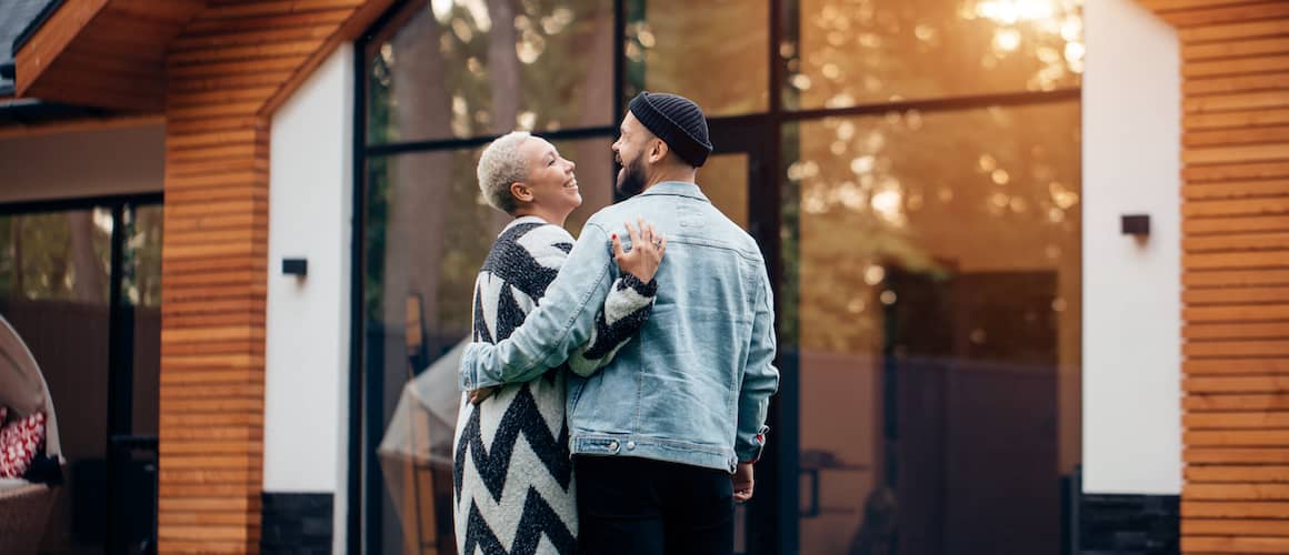 A couple standing outside their house, with a car parked in the driveway.