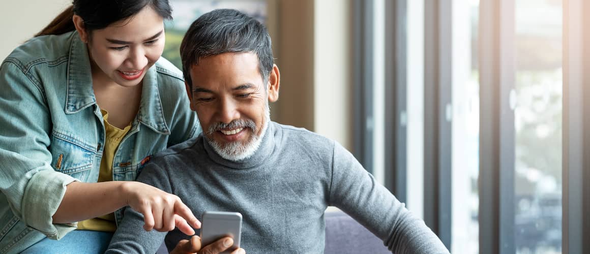 Father and daughter with black hair sitting together while daughter shows father something on a smart phone.