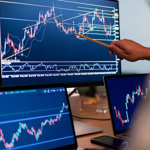 Hand pointing with pencil toward computer monitor showing financial graphs.