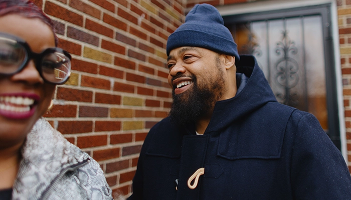 Man in blue beanie smiling at woman in black glasses in front of a brick house.