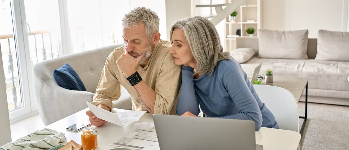 Senior couple sitting together at home in front of a laptop and looking over a paper.