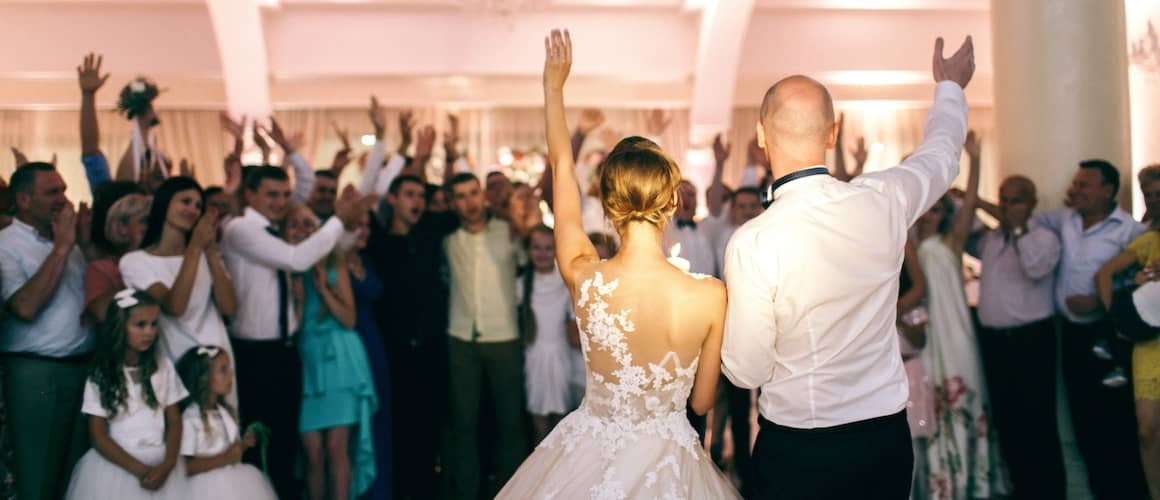 Bride and groom holding hands with arms raised walking into wedding reception filled with standing guests.