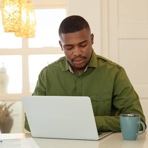 A man in green sitting at a counter with a French press and a laptop.