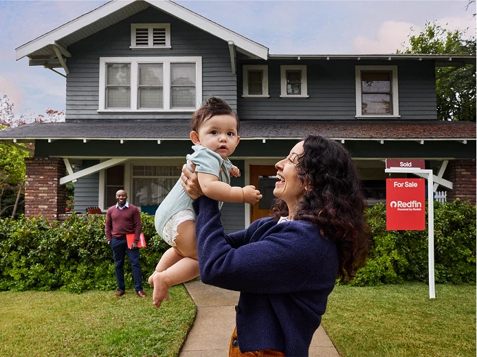 A woman lifting a baby in front of a green two-story house with a Redfin “Sold” sign in the yard while the realtor stands behind them.