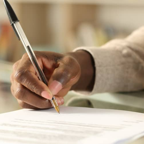 A Black man's hands signing a document on a desk at home, potentially related to paperwork or contracts.
