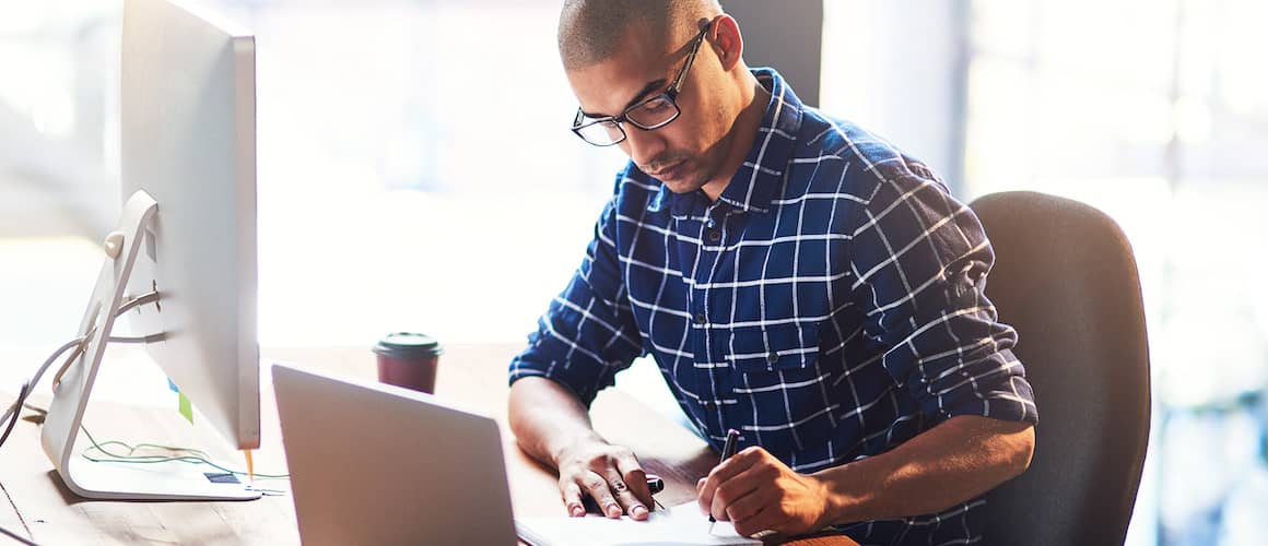 Focused man at desk writing down notes while desktop and laptop.