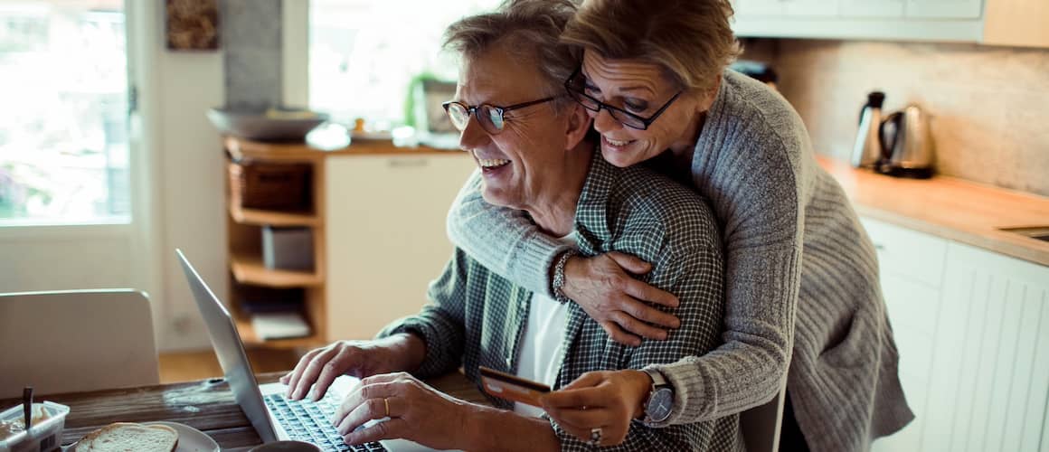 Retired couple in kitchen paying bills online together.