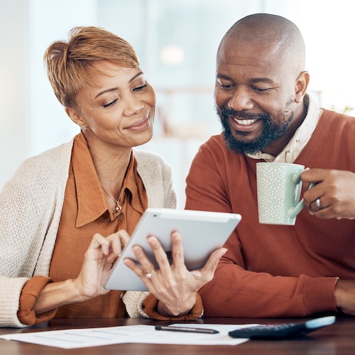 Woman smiling while reviewing finances on tablet while man drinks coffee and looks at the screen. Paperwork and calcluator are on the table nearby.