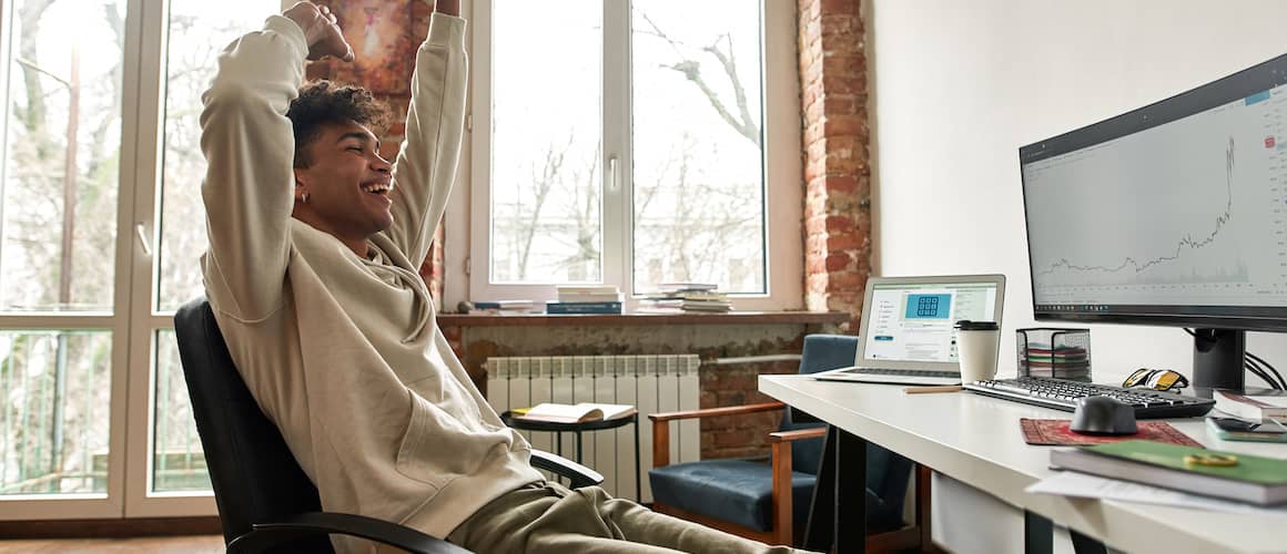 Young trader smiling and hands up after doing well with stocks as seen on desktop.