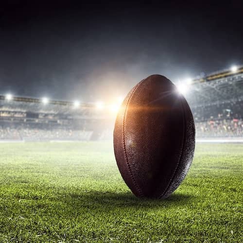 A football at the forefront of a stadium with bright lights on in the background.