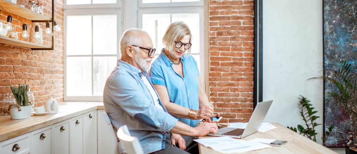 Elderly couple using computer together in bright, sunny kitchen.