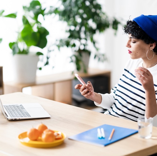 Young woman checking phone at desk with open laptop.