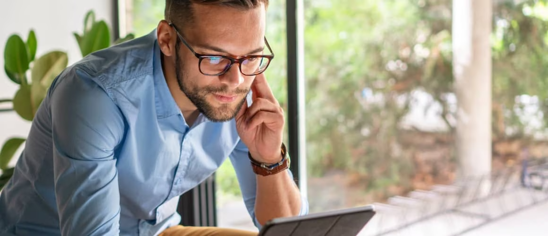 Young man in blue button up shirt leaning on desk and looking at laptop.