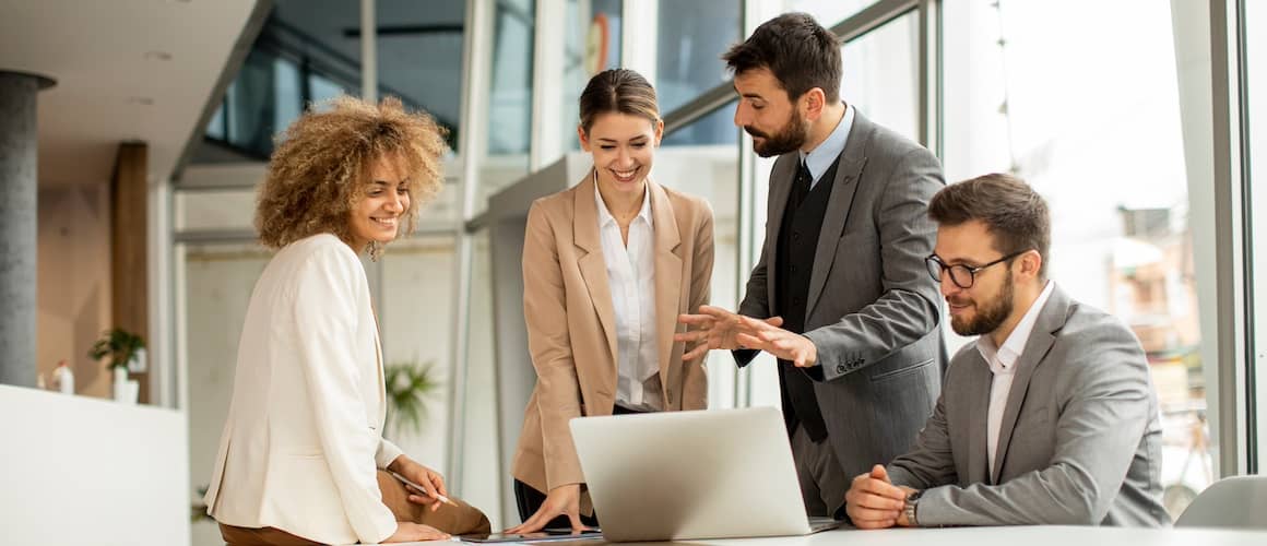 Group of business people gather at a table around an open laptop computer.