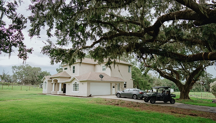 Large beige house on flat green land with cars in driveway.