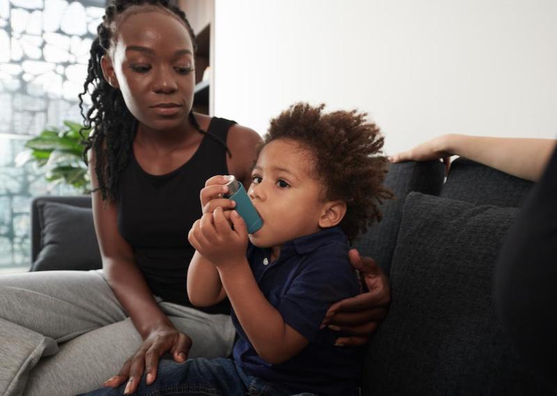 Mother with young son using asthma medication inhaler on couch at home.