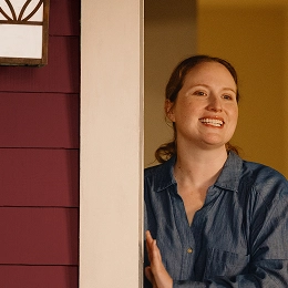 A smiling woman stands in a doorway, partially framed by the door and exterior siding, wearing a denim shirt and looking outward under warm indoor lighting.