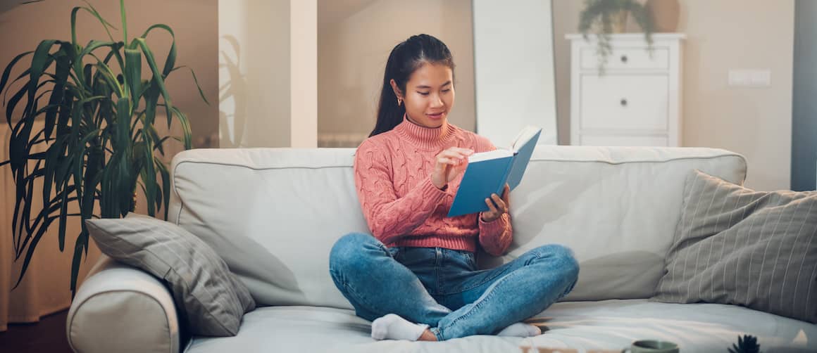 Relaxed brunette woman in sweats curled up reviewing financial paperwork binder on living room sofa.