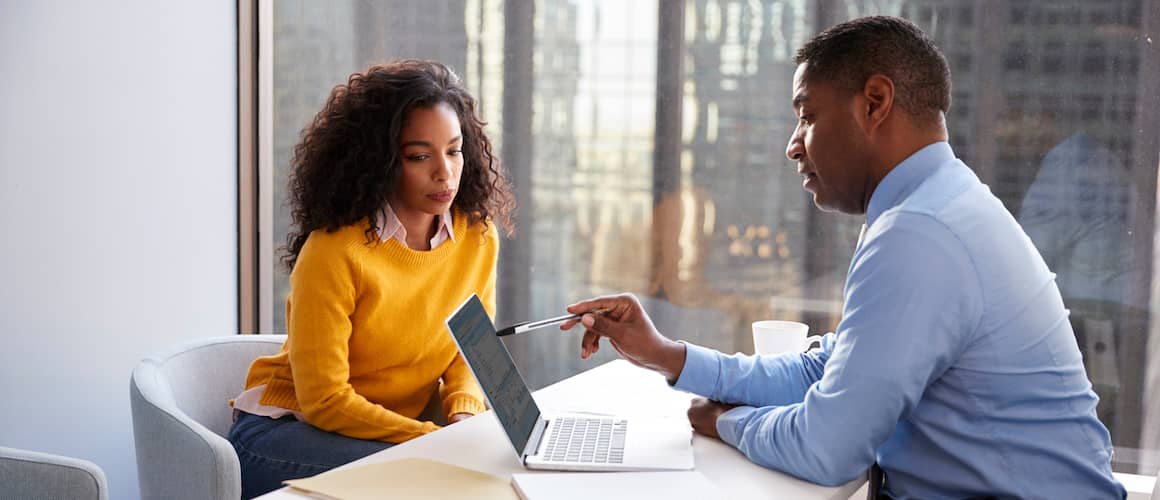 An African American woman in an orange sweater talking to a financial advisor in office.