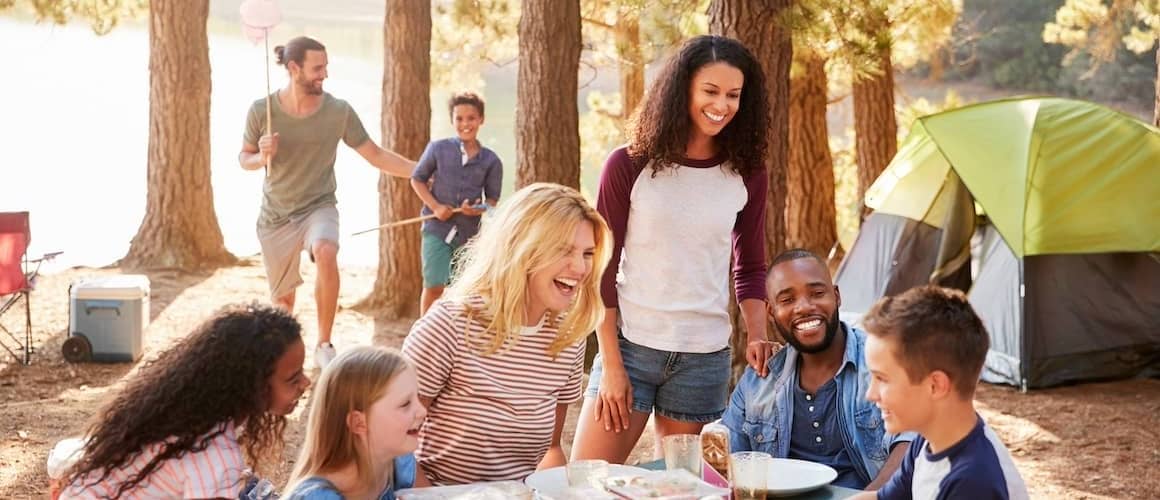 Group of friends with their kids at campsite with trees, a lake, a table and tent.