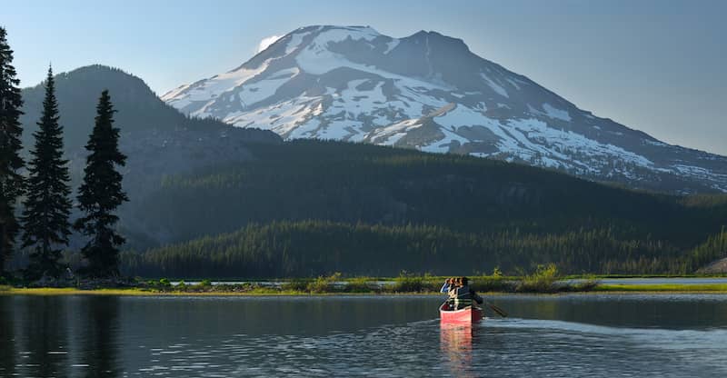 RHB Assets From IGX: A pair of individuals canoeing on a calm lake near Bend, Oregon surrounded by lush green trees and mountains in the distance.