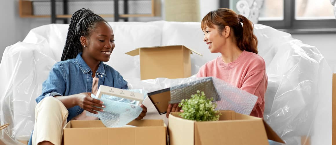 Young female couple unpack boxes together while sitting on the floor of their new home, smiling.