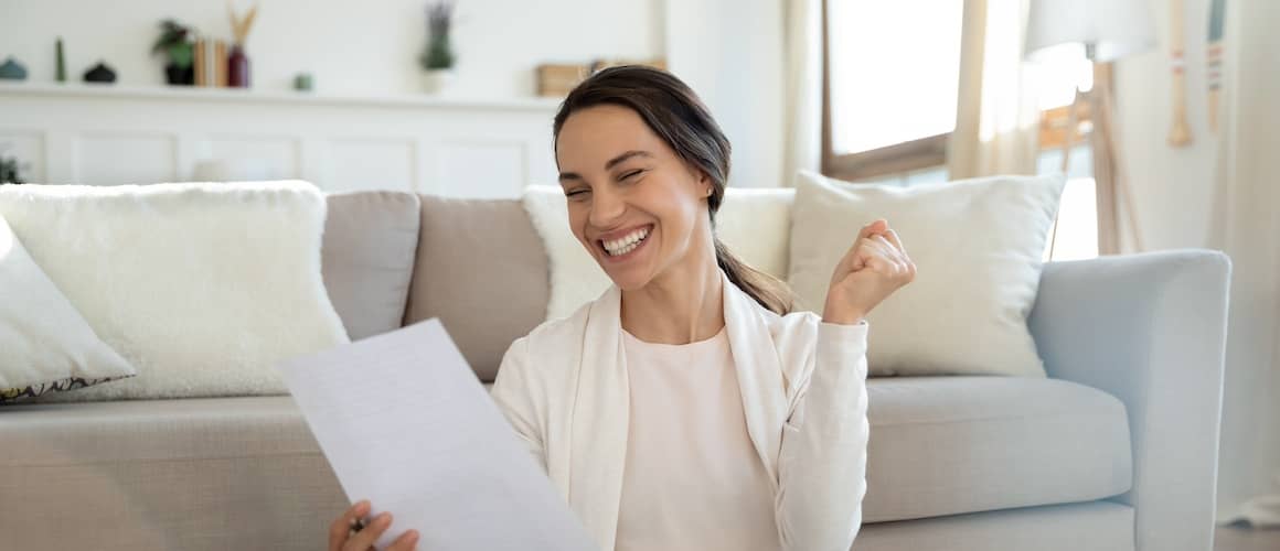 A woman sitting on the floor in front of her couch and smiling at a sheet of paper she's holding.