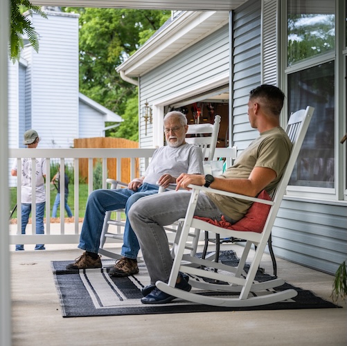 Young man talking to his grandfather, possibly about their military service experiences, while sitting in rocking chairs on the porch.
