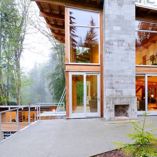 Contemporary home on pine tree wooded lot featuring mirrored picture windows and stone outdoor fireplace.