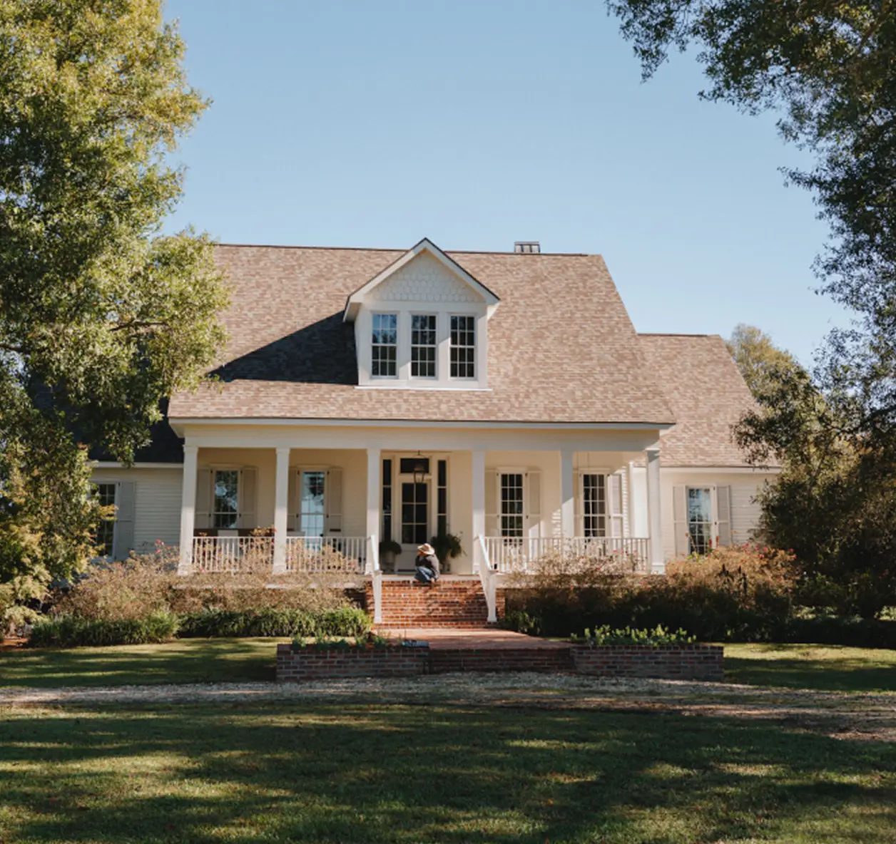 A two-story white house with a large front porch, dormer windows, and landscaped yard, framed by trees under a clear blue sky.