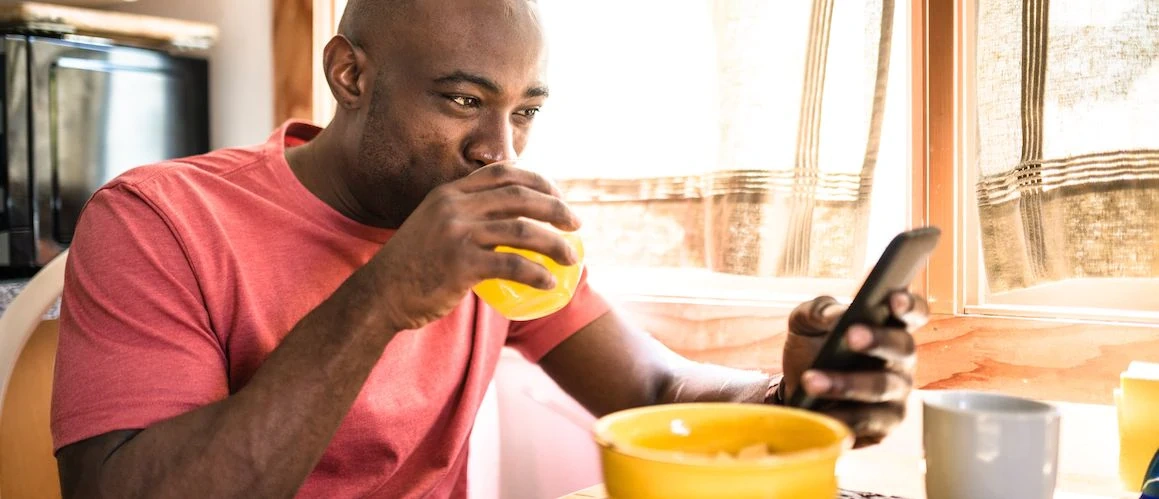 A black man having breakfast. He is drinking a glass of orange juice, with a bowl of cereal in front of him. He is looking at a smart phone that is in his left hand.