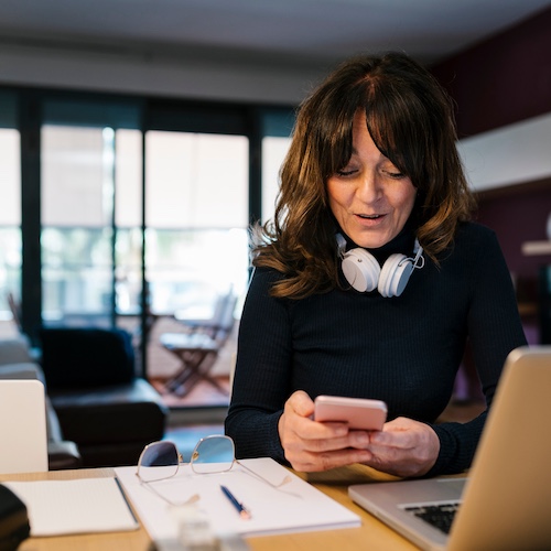 Woman with headphones on neck, working on phone, with laptop open and paperwork nearby in her home office.