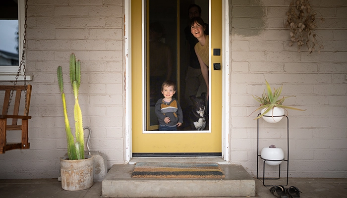 Small child, dog, mother, and father looking out front porch door smiling.