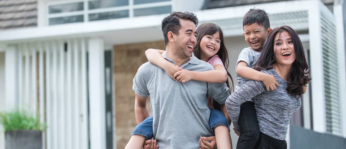 A couple with two kids giving piggyback rides in front of their house, symbolizing family happiness and homeownership.