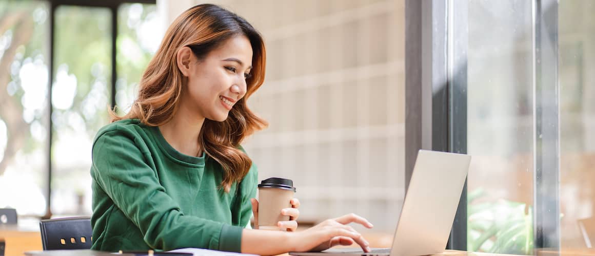 Smiling Asian businesswoman using her laptop while holding a cup of coffee.