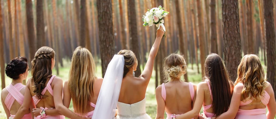 View from behind of a bride and her bridesmaids embracing one another looking at a forest.