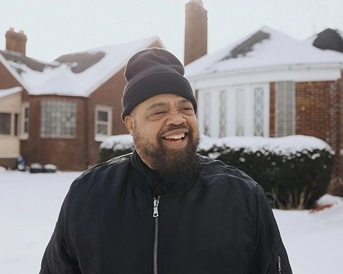 Man standing in the snow in front of brick house wearing black beanie and black zip up coat.