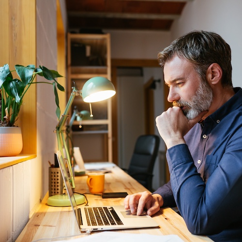 Man at home office desk looking at laptop with a pensive expression.