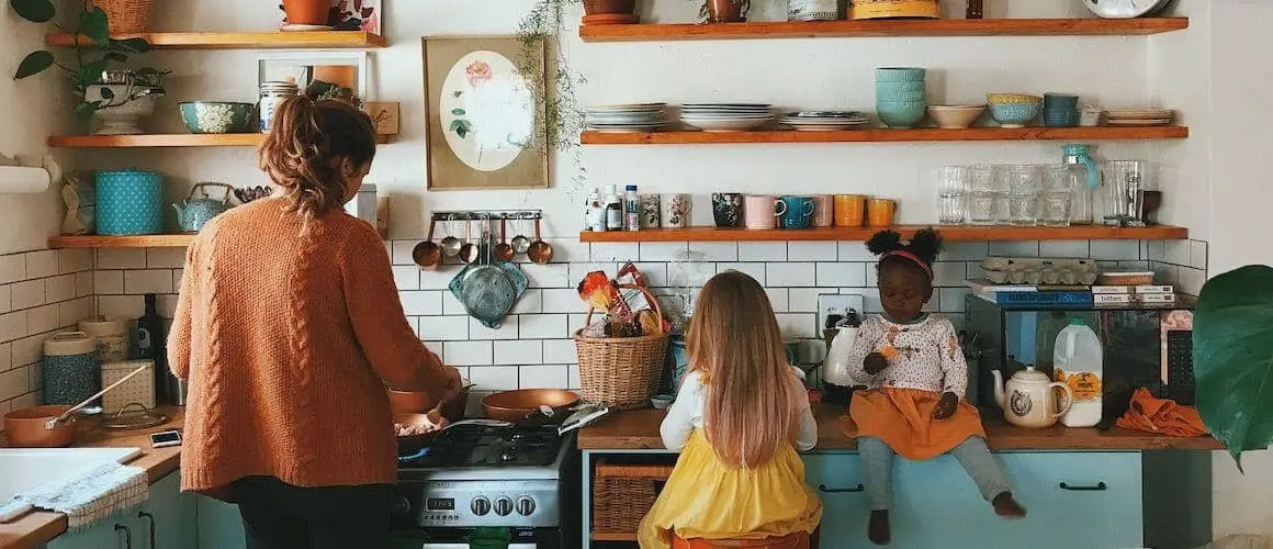 A mom cooks food in on the stove while her two daughters eat nearby.