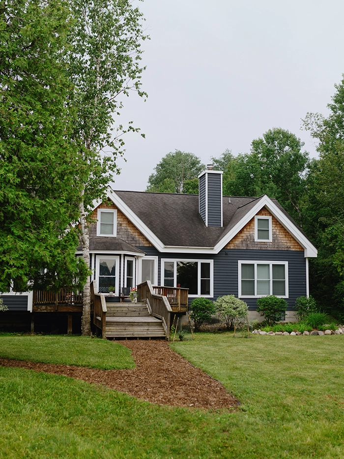 Dark blue house with a front deck and stairs, surrounded by trees and greenery.