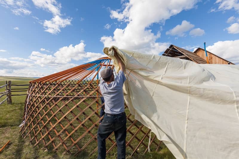 Man constructing simple and small yurt pulling fabric over latticed structure.