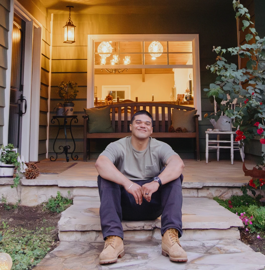 A young man sits smiling on the front steps of a home in the evening, with a warmly lit porch behind them.