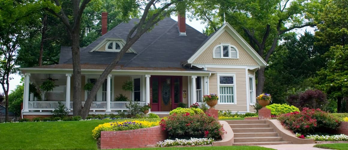 Tan house with a double-layered front lawn, brick steps, and a wrap-around porch.