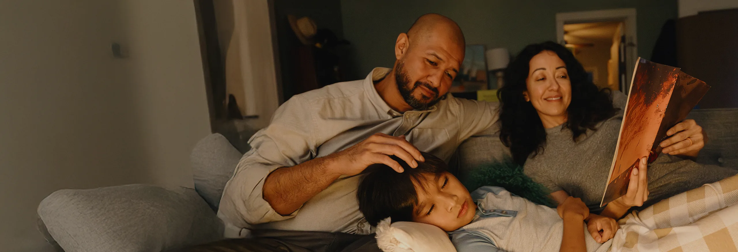 Two parents sitting on a couch with a child resting on a pillow, while the mother reads a book.
