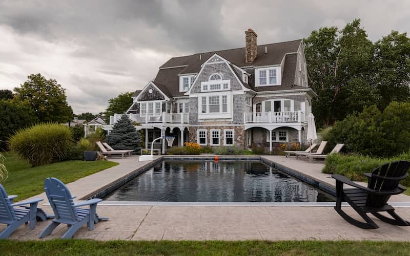 Large Craftsman home in gray and stone with a pool surrounded by lounge chairs.