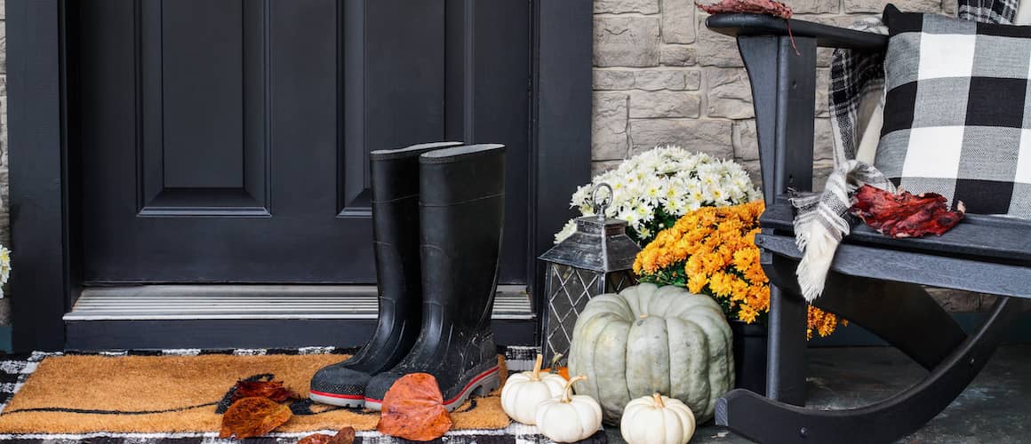 Black door with black rain boots out front, small white pumpkins, white mums and a rocking chair to the side with a black and white plaid blanket strewn over it.