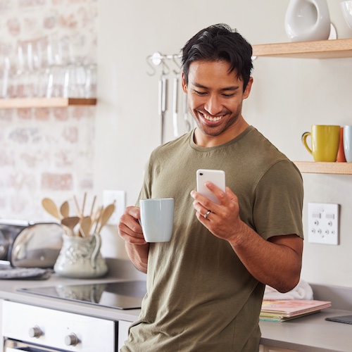 Man in kitchen drinking coffee and smiling while looking at smart phone.