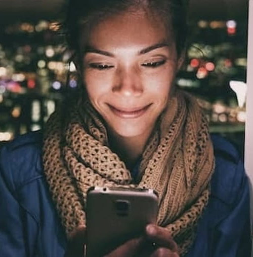 A woman standing in front of a window, engrossed in her phone.