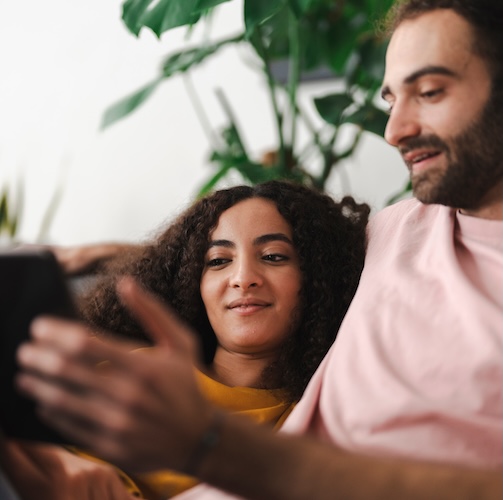 A couple sitting together on a couch, looking at a tablet in a bright and modern living room filled with greenery, enjoying a cozy and relaxed moment at home.
