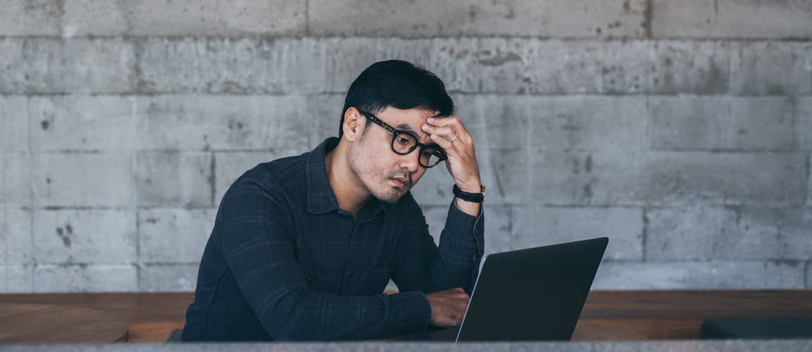 A young Asian man looking stressed while using a laptop, possibly in a home or work setting.